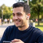 Dışarıda yürüyüş yaparken kulağında yeni nesil kablosuz bluetooth kulaklık takılı olan ve akıllı telefonunu kullanan mutlu erkek kullanıcı.